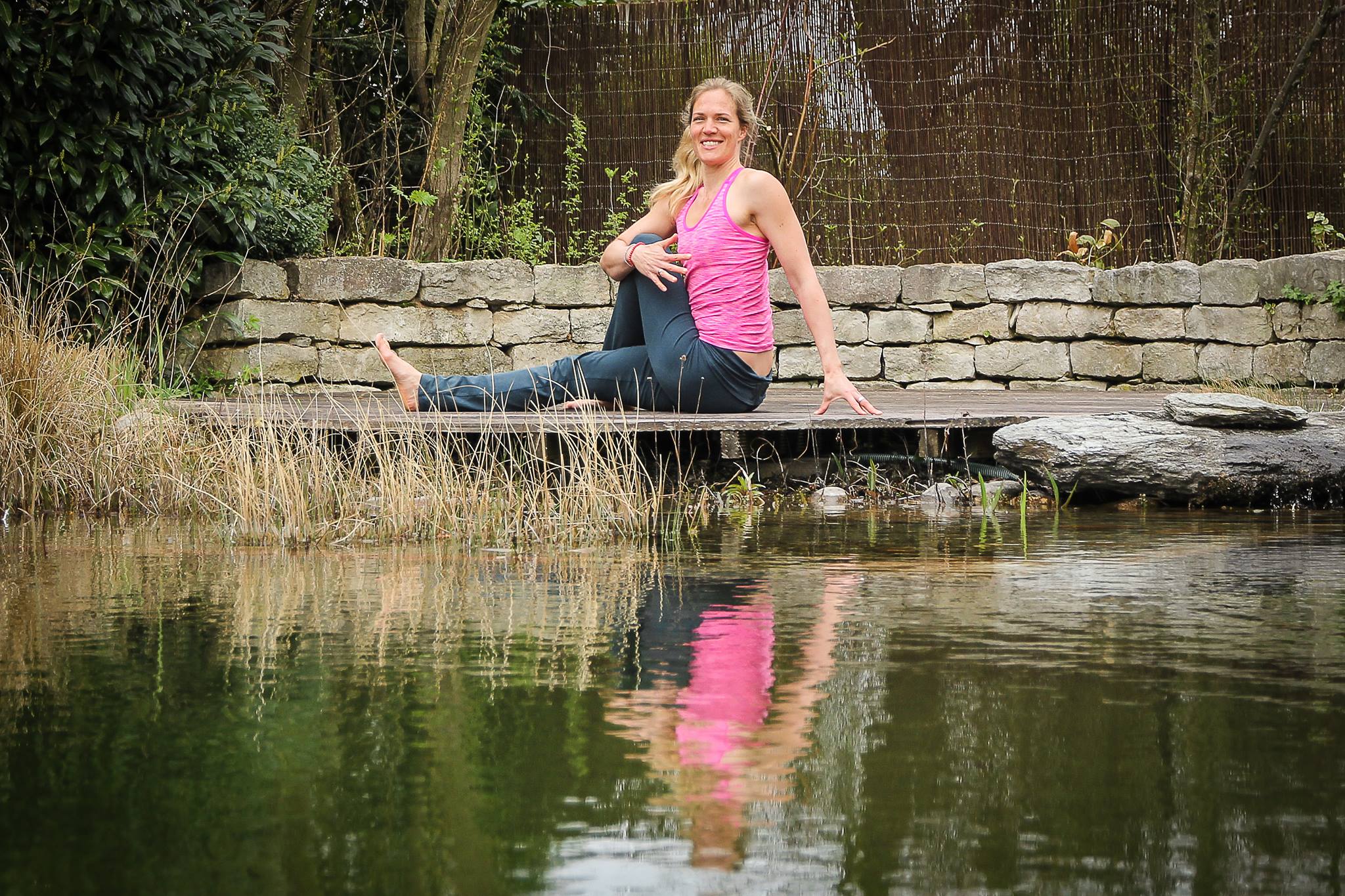 Frau in rosa Tanktop und blauer Yoga-Hose sitzt lächelnd in einer Yoga-Pose (Drehsitz) auf einer Holzplattform am Rand eines Teichs, umgeben von Gräsern, einer Steinmauer und Bäumen.